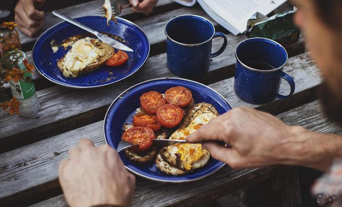 Image taken from above of a couple eating breakfast outdoors. Two plates of fried eggs and tomatoes on toast with cups of hot coffee are visible.
